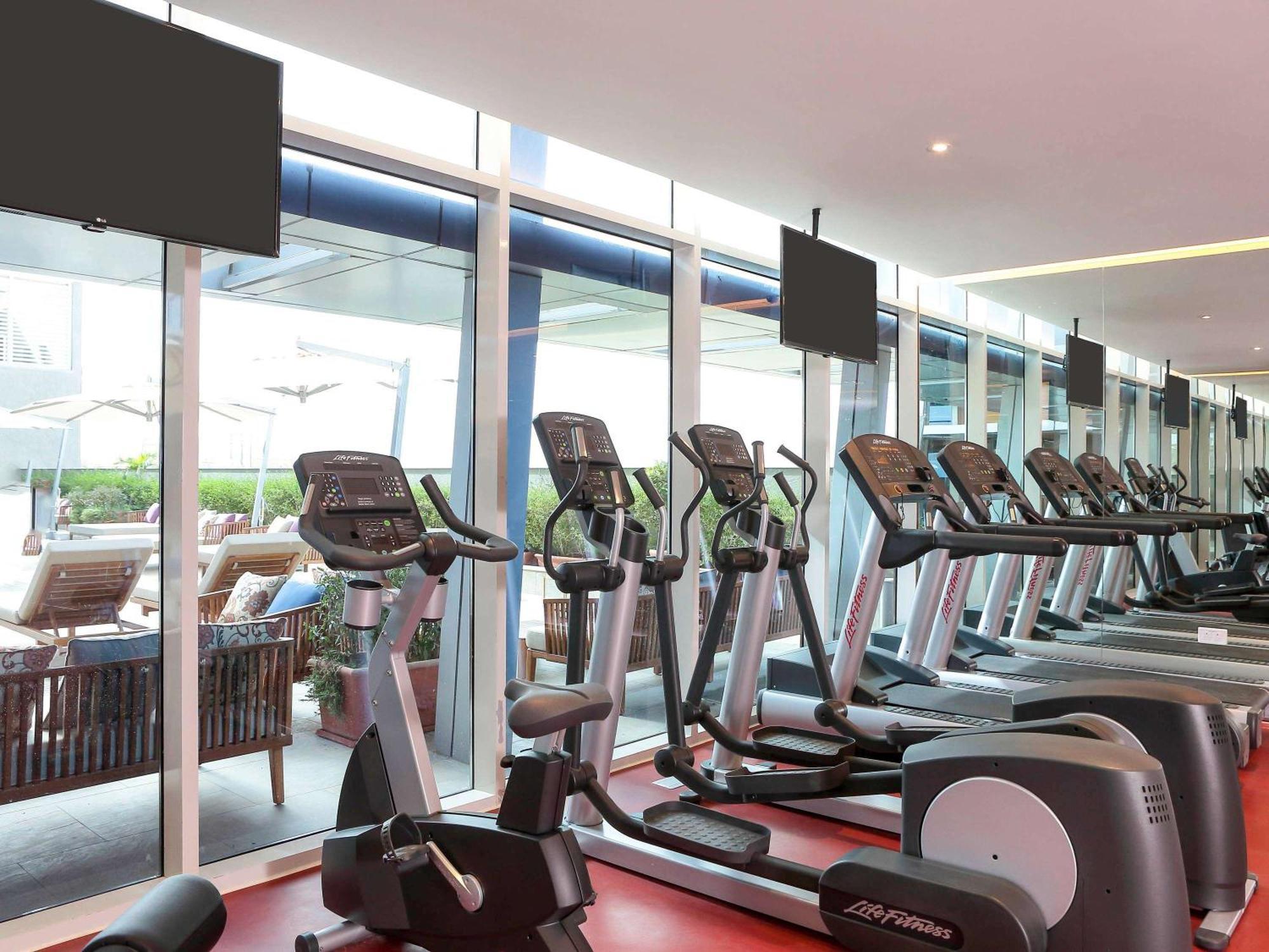 Novotel Jazan מראה חיצוני תמונה The photo shows a modern gym interior with several exercise machines, specifically elliptical trainers, arranged neatly in rows. Large windows allow natural light to fill the space, providing a view outside to a patio area with lounge chairs. There a